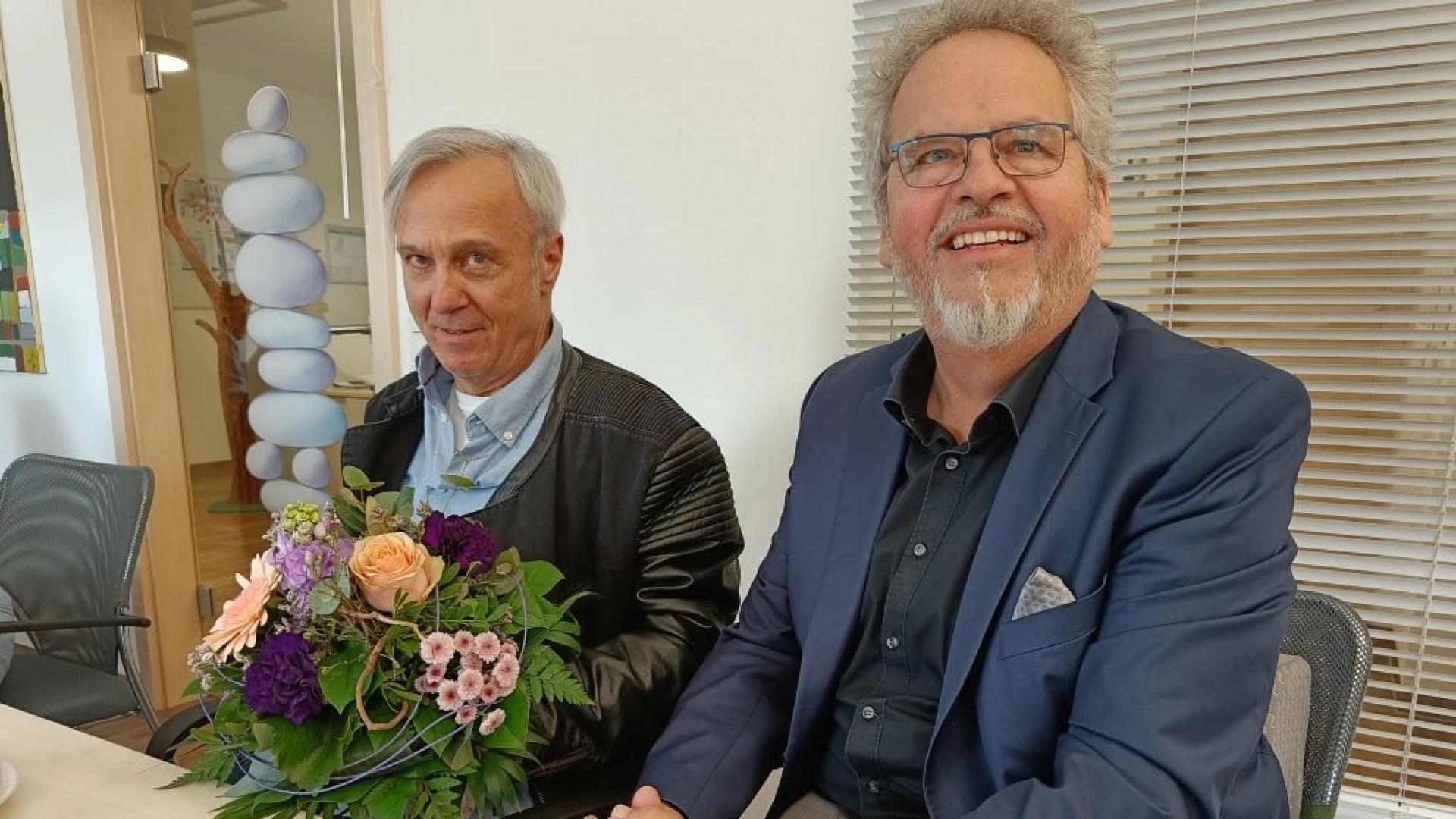 Zwei M&auml;nner sitzen nebeneinander an einem Tisch und l&auml;cheln in die Kamera. Der Mann links h&auml;lt einen Blumenstrau&szlig; in den H&auml;nden, er tr&auml;gt eine schwarze Lederjacke. Der Mann rechts tr&auml;gt ein blaues Jackett, drunter ein dunkles Hemd und eine Brille.