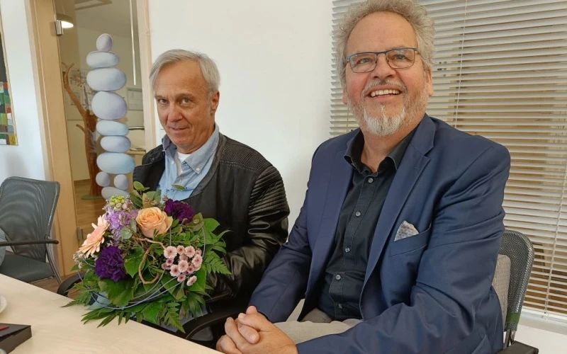 Zwei M&auml;nner sitzen nebeneinander an einem Tisch und l&auml;cheln in die Kamera. Der Mann links h&auml;lt einen Blumenstrau&szlig; in den H&auml;nden, er tr&auml;gt eine schwarze Lederjacke. Der Mann rechts tr&auml;gt ein blaues Jackett, drunter ein dunkles Hemd und eine Brille.