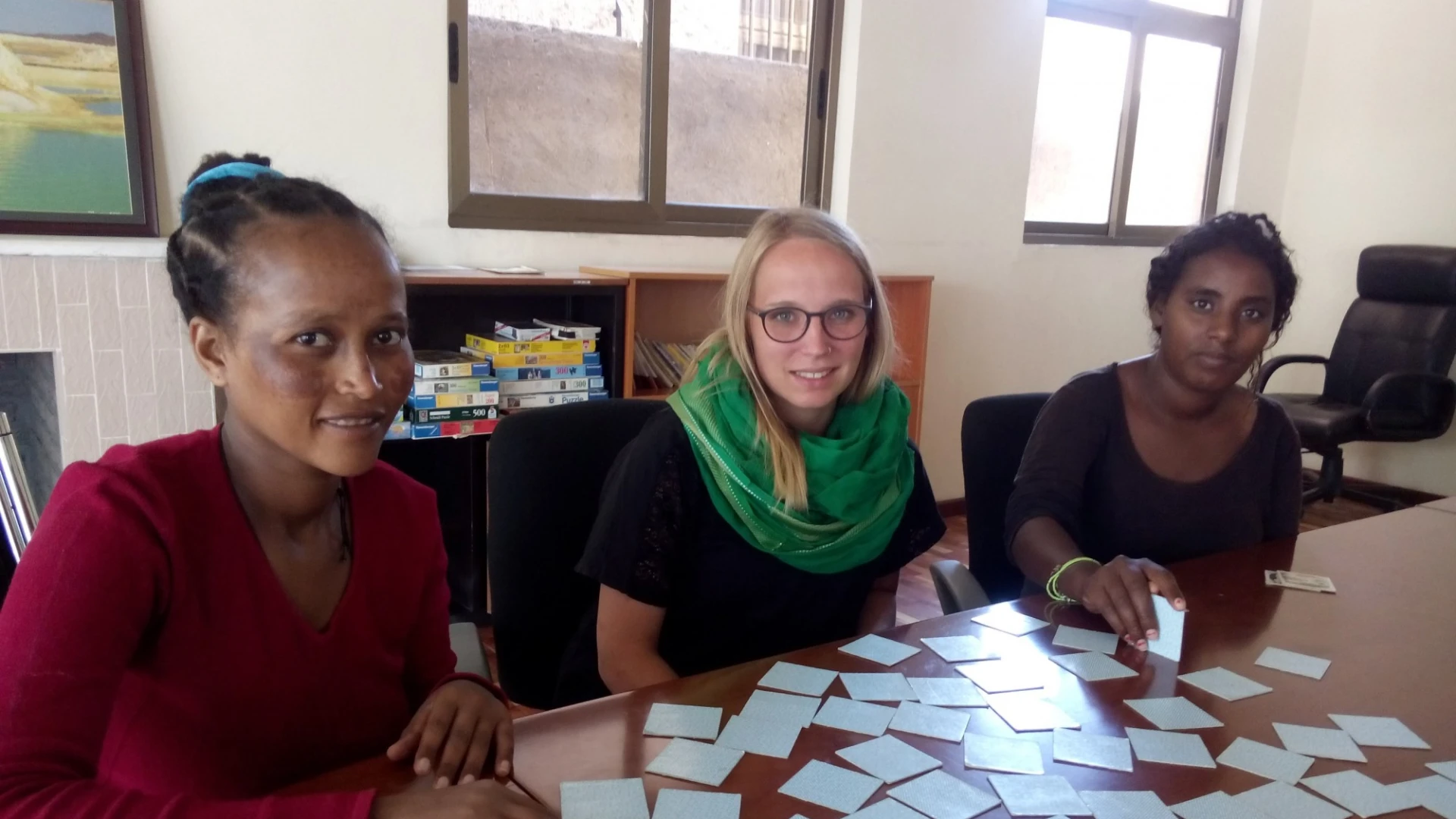 Drei Frauen sitzen an einem Tisch. Sie spielen Memory. Im Hintergrund sind zwei Fenster, ein Stuhl und ein Regal zu sehen.