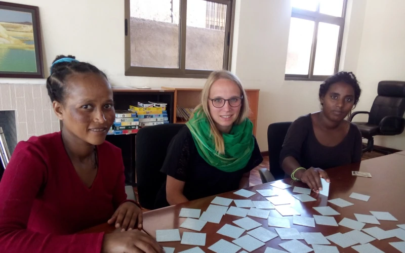 Drei Frauen sitzen an einem Tisch. Sie spielen Memory. Im Hintergrund sind zwei Fenster, ein Stuhl und ein Regal zu sehen.