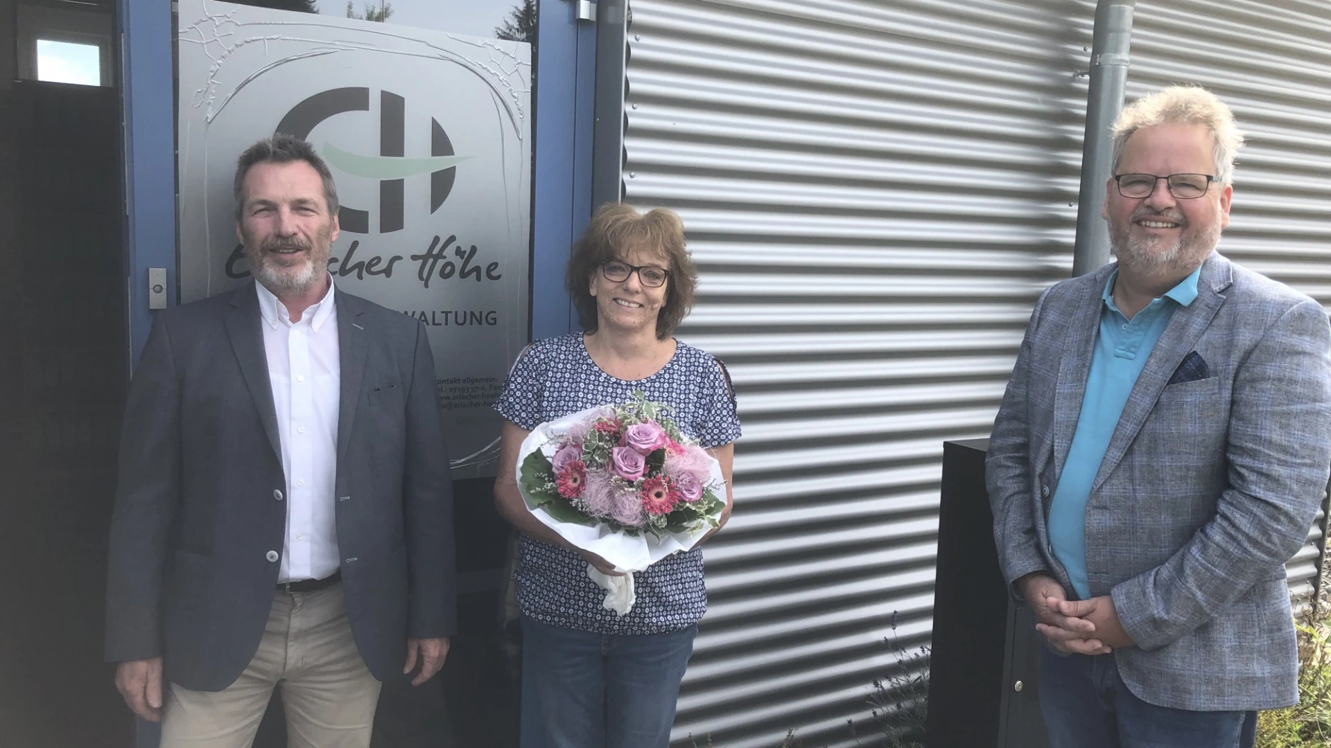 Drei Personen stehen vor einer T&uuml;r mit dem Logo der Erlacher H&ouml;he. Die Frau in der Mitte h&auml;lt einen Blumenstrau&szlig; in der Hand. Alle drei l&auml;cheln in die Kamera.