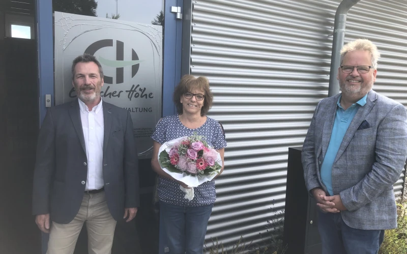 Drei Personen stehen vor einer T&uuml;r mit dem Logo der Erlacher H&ouml;he. Die Frau in der Mitte h&auml;lt einen Blumenstrau&szlig; in der Hand. Alle drei l&auml;cheln in die Kamera.