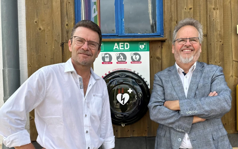 Zwei M&auml;nner stehen vor einem Defibrillators an einem Geb&auml;ude und l&auml;cheln in die Kamera.