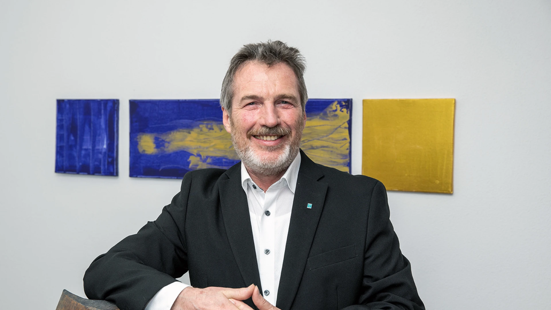 L&auml;chelnder Mann mittleren Alters mit grauem Haar und Bart in einem schwarzen Jackett vor bunten Kunstwerken, die H&auml;nde gefaltet..