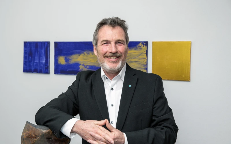 L&auml;chelnder Mann mittleren Alters mit grauem Haar und Bart in einem schwarzen Jackett vor bunten Kunstwerken, die H&auml;nde gefaltet..