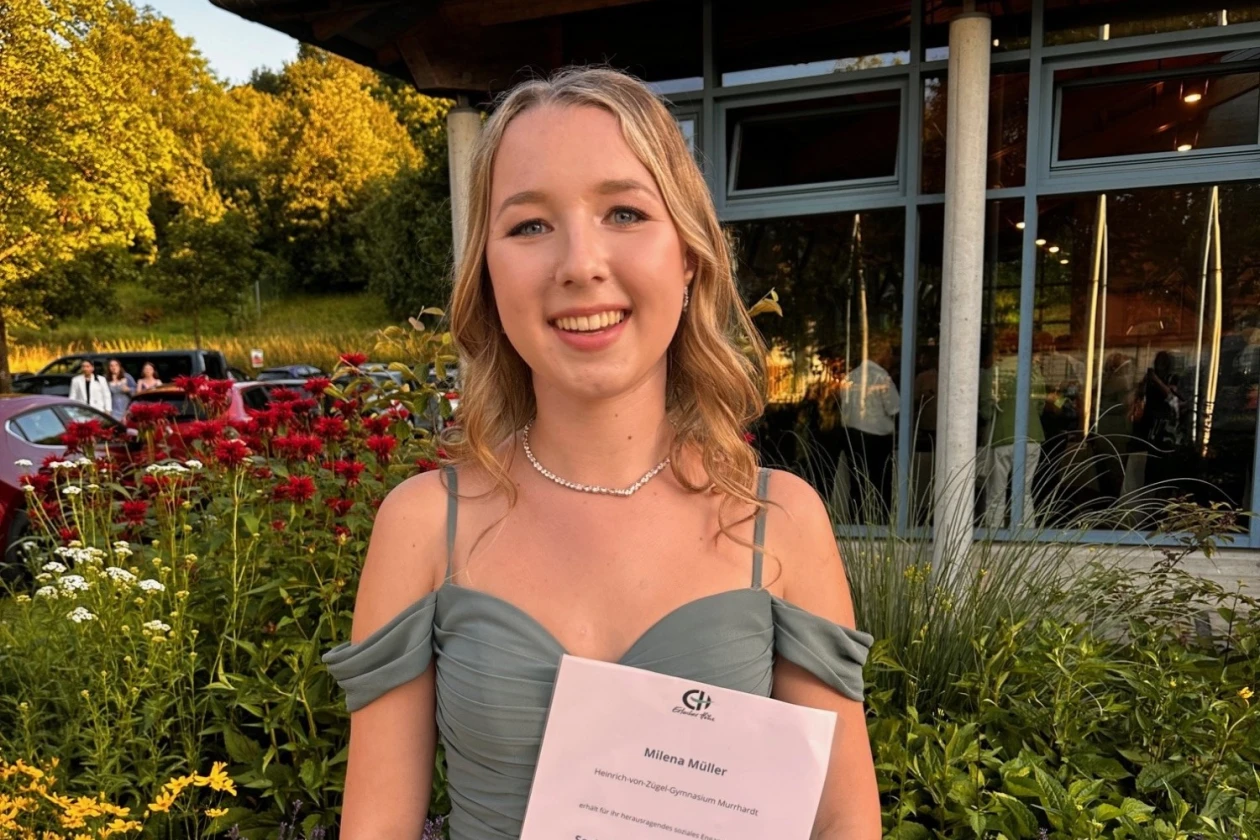 Eine junge blondhaarige Frau h&auml;lt vor einem &ouml;ffentlichen Geb&auml;ude ihre Urkunde f&uuml;r den Sozialpreis in der Hand. Sie l&auml;chelt in die Kamera und tr&auml;gt ein festliches, olivgr&uuml;nes Kleid.