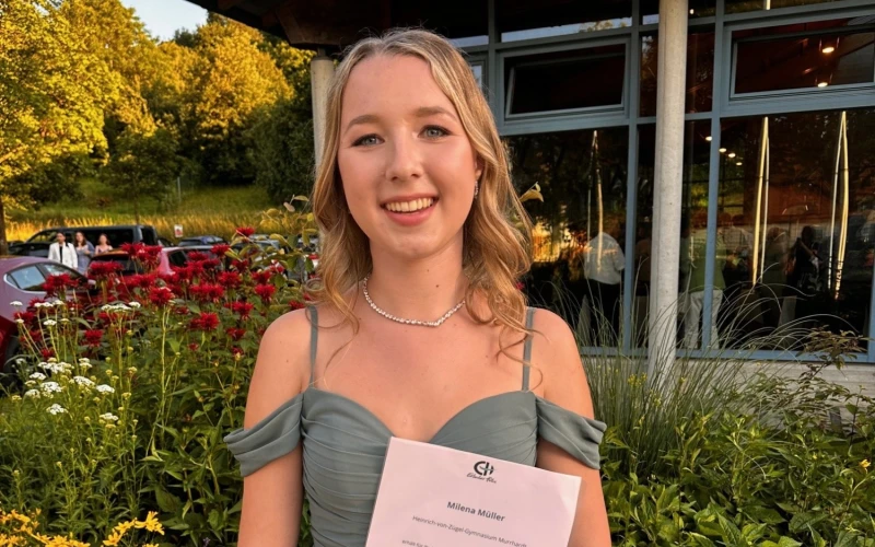 Eine junge blondhaarige Frau h&auml;lt vor einem &ouml;ffentlichen Geb&auml;ude ihre Urkunde f&uuml;r den Sozialpreis in der Hand. Sie l&auml;chelt in die Kamera und tr&auml;gt ein festliches, olivgr&uuml;nes Kleid.