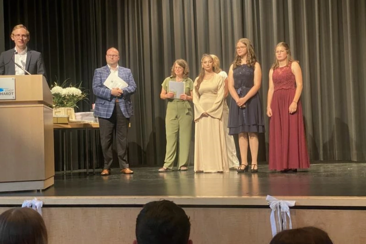 Ein Junger Mann mit Sakko steht am Rednerpult. Junge Menschen in festlicher Kleidung stehen im Hintergrund. Sie h&ouml;ren aufmerksam zu.
