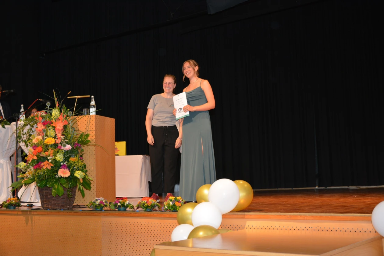Eine junge Frau in langem Abendkleid mit einer Urkunde in der Hand l&auml;chelt mit einer Frau im T-Shirt mit Logo der Erlacher H&ouml;he in die Kamera. Beide stehen auf der B&uuml;hne einer Festhalle, die festlich geschm&uuml;ckt ist.