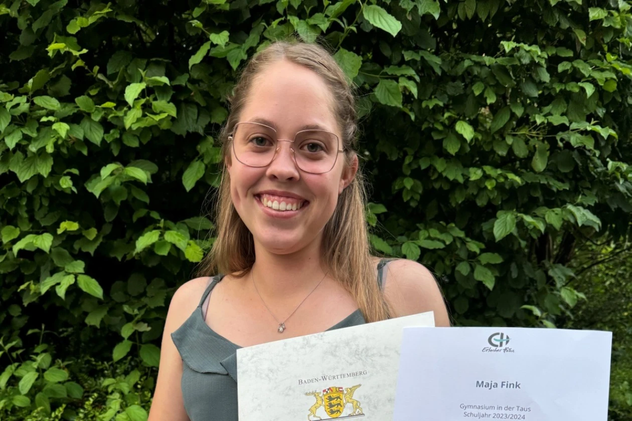 Eine l&auml;chelnde junge blondhaarige Frau mit Brille in festlichem Outfit pr&auml;sentiert ihr Zeugnis und Ihre Urkunde f&uuml;r den Sozialpreis. Sie steht vor einer dichten gr&uuml;nen Hecke.