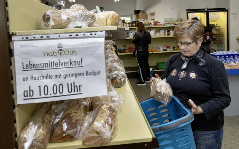Eine Frau mit blauem Einkaufskorb steht vor einem Regal mit verpackten Backwaren in einem Lebensmittelgesch&auml;ft. Ein Schild weist auf den Lebensmittelverkauf f&uuml;r Haushalte mit geringem Budget ab 10.00 Uhr hin. Im Hintergrund ist eine weitere Person an Lebensmittelregalen zu sehen.