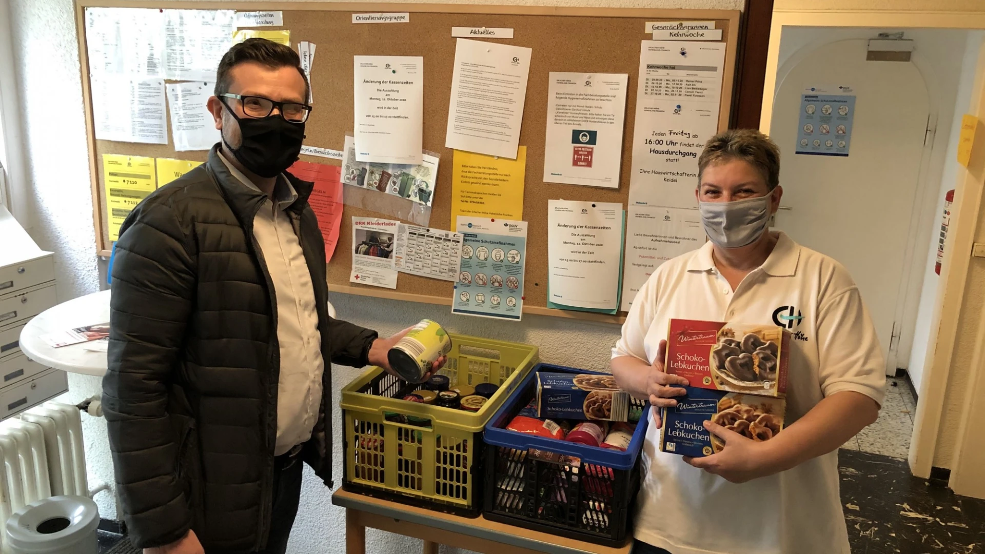 Zwei Personen stehen in einem Raum mit zwei Kisten Lebensmittel. Beide tragen einen Mund-Nasenschutz. Der Mann links h&auml;lt eine Dose in der Hand und die Frau rechts zwei Packungen Lebkuchen. Im Hintergrund ist eine Pinnwand mit Dokumenten und ein Stehtisch zu sehen.