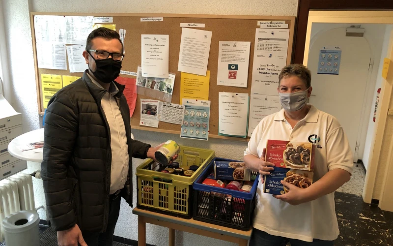 Zwei Personen stehen in einem Raum mit zwei Kisten Lebensmittel. Beide tragen einen Mund-Nasenschutz. Der Mann links h&auml;lt eine Dose in der Hand und die Frau rechts zwei Packungen Lebkuchen. Im Hintergrund ist eine Pinnwand mit Dokumenten und ein Stehtisch zu sehen.