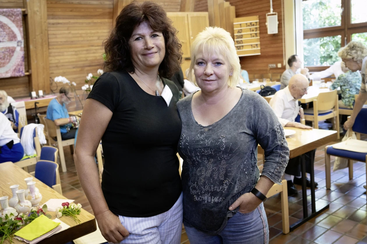 Zwei Frauen stehen nebeneinander in einem Raum mit Holzw&auml;nden, eingedeckten Tischen und St&uuml;hlen. Die Frau links tr&auml;gt ein schwarzes T-Shirt, die Frau rechts ein graues Oberteil. Sie l&auml;cheln in die Kamera, im Hintergrund sind weitere Personen zu sehen.