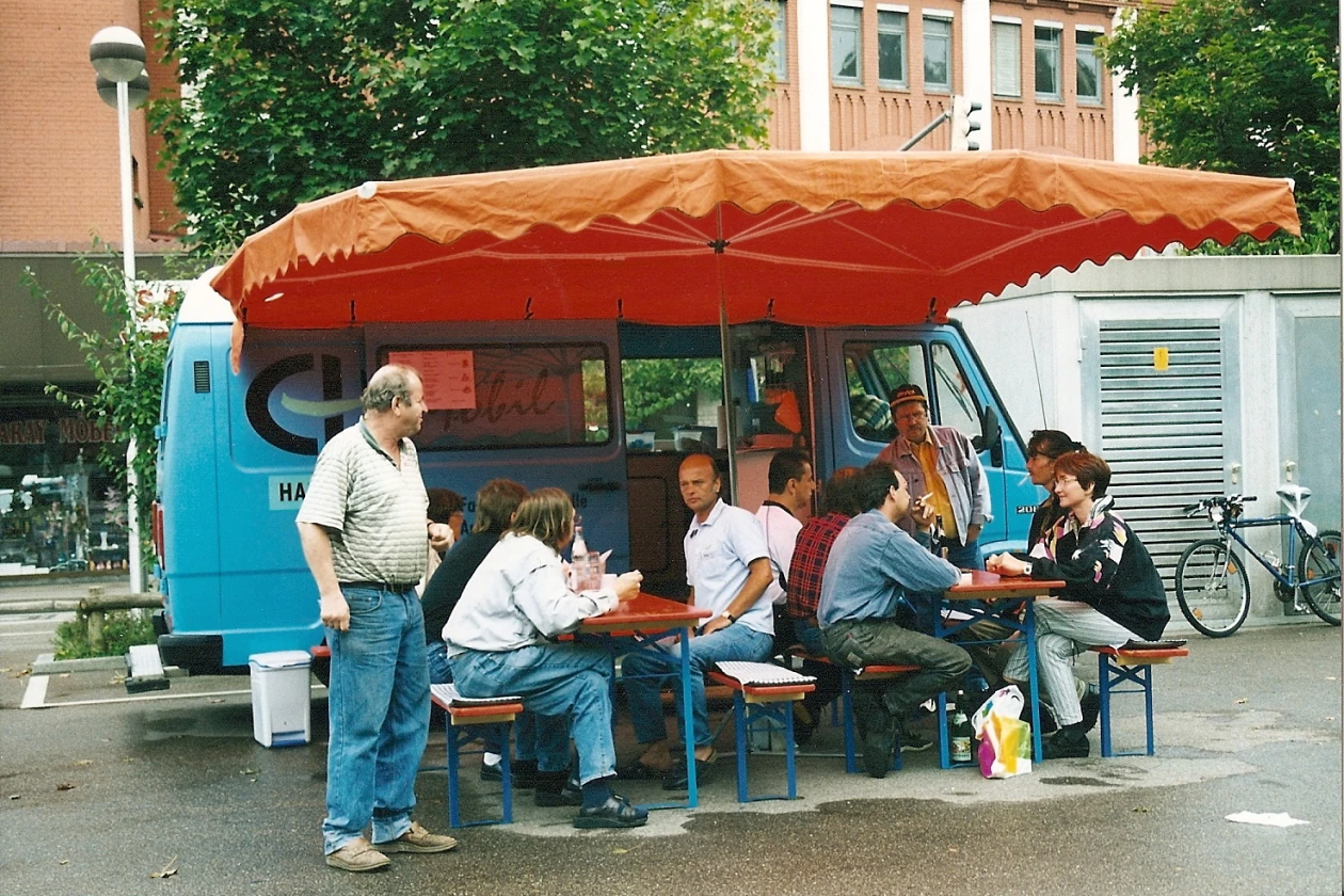 Blaues EH mobil mit rotem Sonnenschirm, unter dem mehrere Menschen an Tischen sitzen und sich unterhalten.