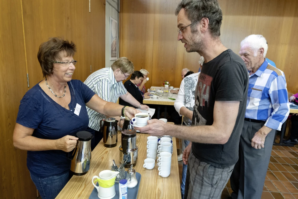 Eine Frau und ein Mann stehen an einer Theke, auf der Kaffeekannen, Tassen und Milch und Zucker stehen. Die Fraureicht dem Mann eine Tasse mit Kaffee. Im Hintergrund sind weitere Personen an Tischen sitzend zu sehen.