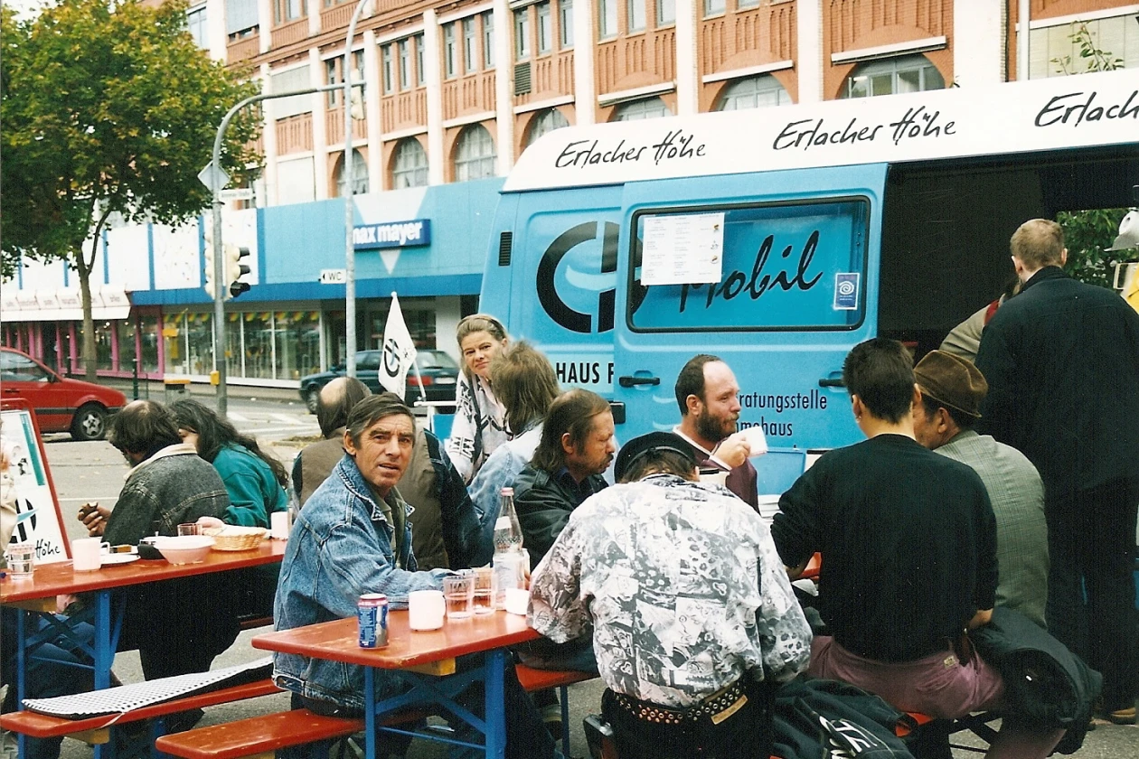 Historische Aufnahme: Menschen an Biertischb&auml;nken mit Getr&auml;nken in einer Stadt vor einem hellblauen Lieferwagen mit der Aufschrift "EH mobil".