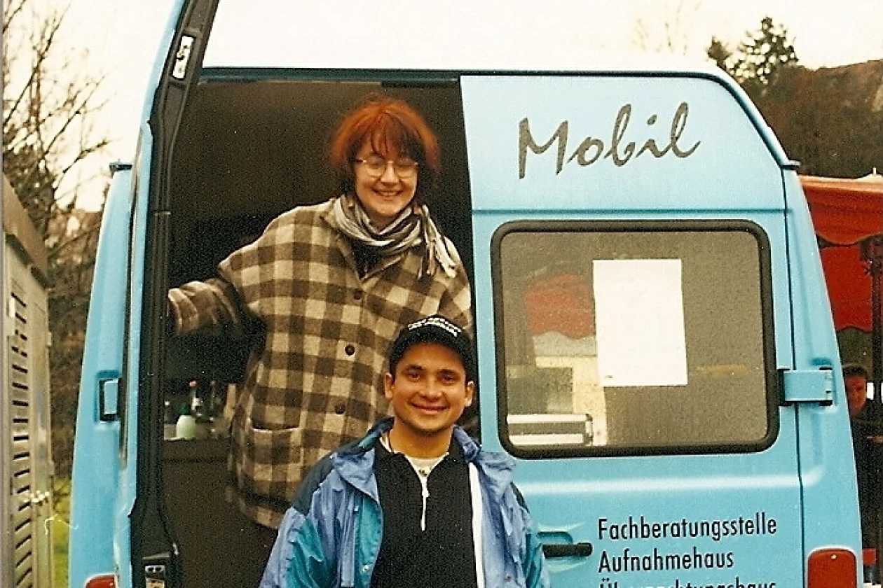 Zwei Personen stehen an einem hellblauen Transporter mit der Aufschrift "Fachberatungsstelle", "Aufnahmehaus", "&Uuml;bernachtungshaus", "Wohnraumhilfen" und "Stra&szlig;ensozialarbeit" steht. Eine Frau in karierter Jacke steht im Ladebereich, ein Mann in blauer Jacke lehnt sich am Heck des Fahrzeugs.