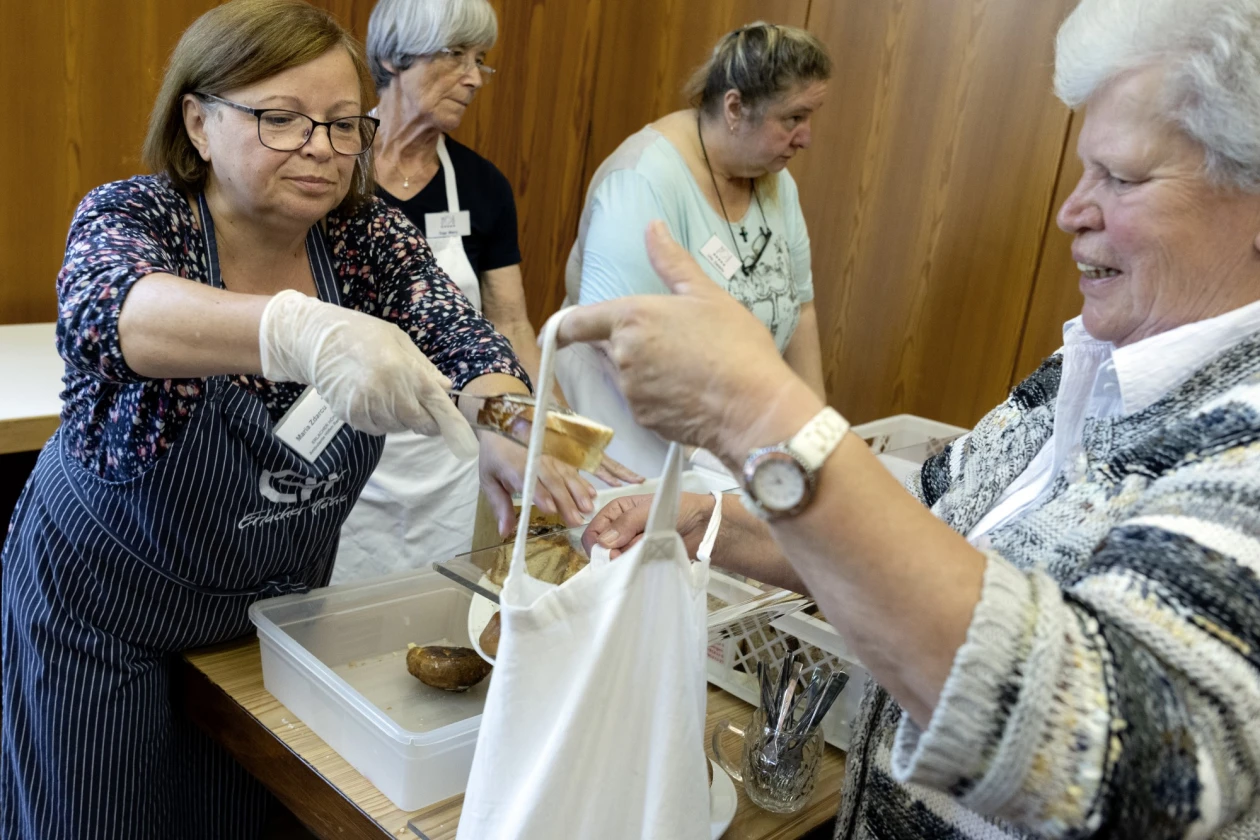 Drei Frauen stehen hinter der Theke. Zwei von ihnen tragen eine Sch&uuml;rze und, eine h&auml;lt mit einer Zange Geb&auml;ck, das sie der Frau vor der Theke in die ge&ouml;ffnete Tragetasche packt.