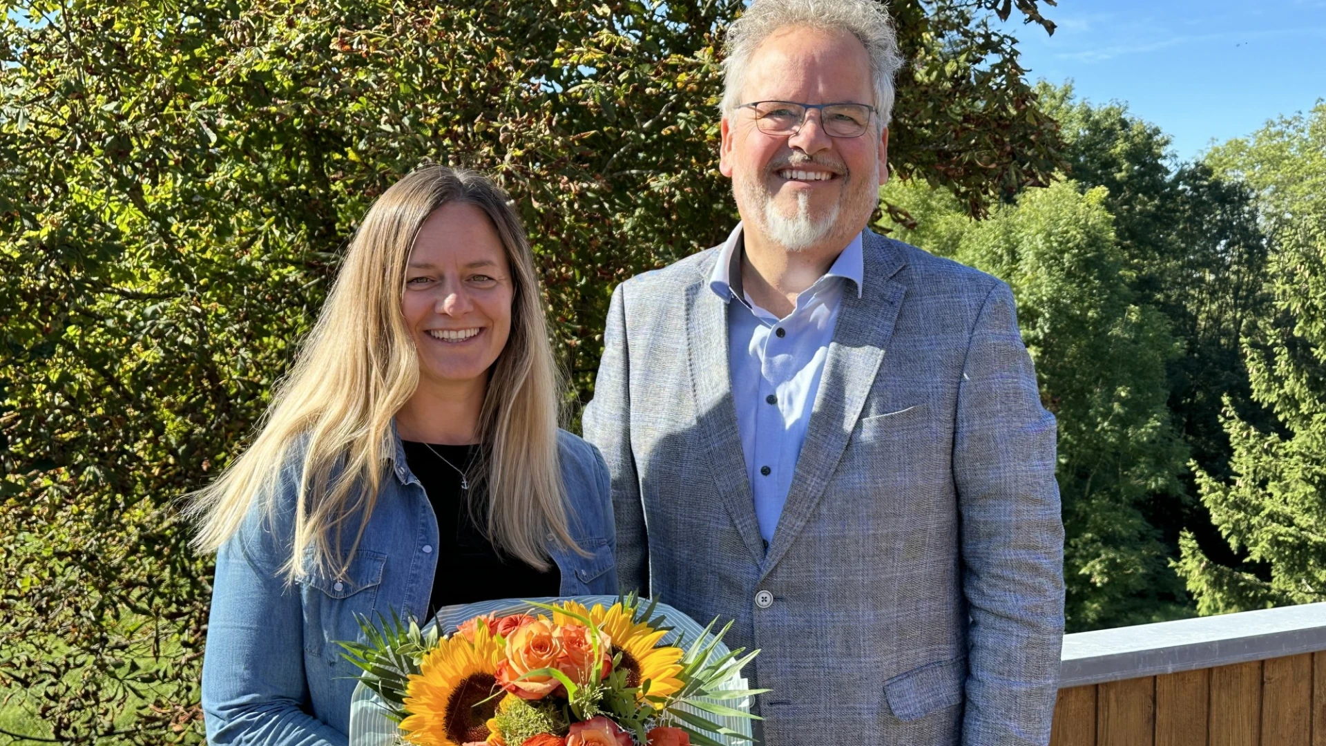 Eine Frau und ein Mann stehen nebeneinander und l&auml;cheln in die Kamera. Die Frau h&auml;lt einen Blumenstrau&szlig; in den H&auml;nden. Der Mann tr&auml;gt eine Brille und ein graues Jackett &uuml;ber einem hellblauen Hemd. Im Hintergrund sind B&auml;ume zu sehen.