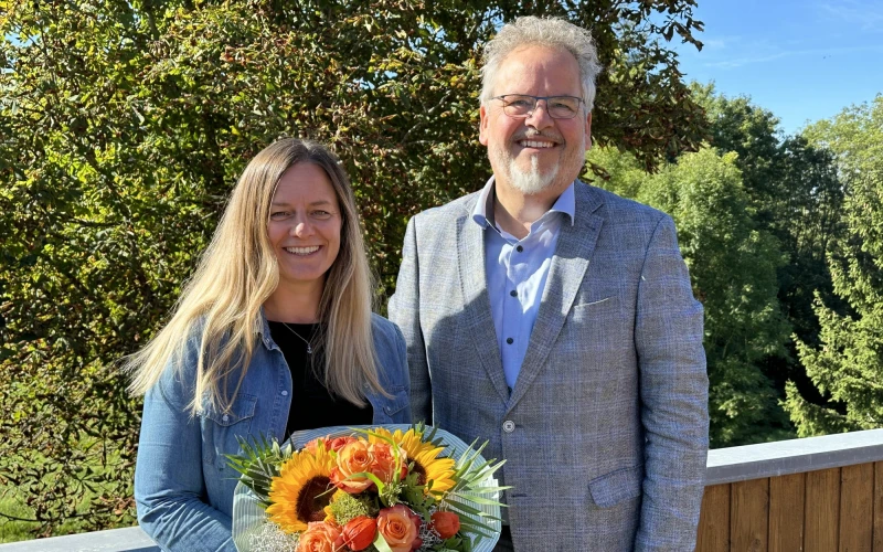 Eine Frau und ein Mann stehen nebeneinander und l&auml;cheln in die Kamera. Die Frau h&auml;lt einen Blumenstrau&szlig; in den H&auml;nden. Der Mann tr&auml;gt eine Brille und ein graues Jackett &uuml;ber einem hellblauen Hemd. Im Hintergrund sind B&auml;ume zu sehen.