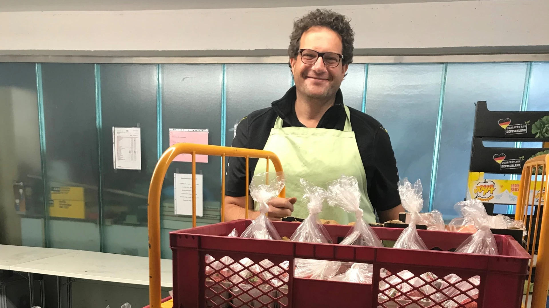 Ein Mann mit lockigen Haaren und Brille tr&auml;gt eine helle Sch&uuml;rze und steht hinter einem Wagen mit mehreren Kisten voller in Folie verpackter Lebensmittelportionen.