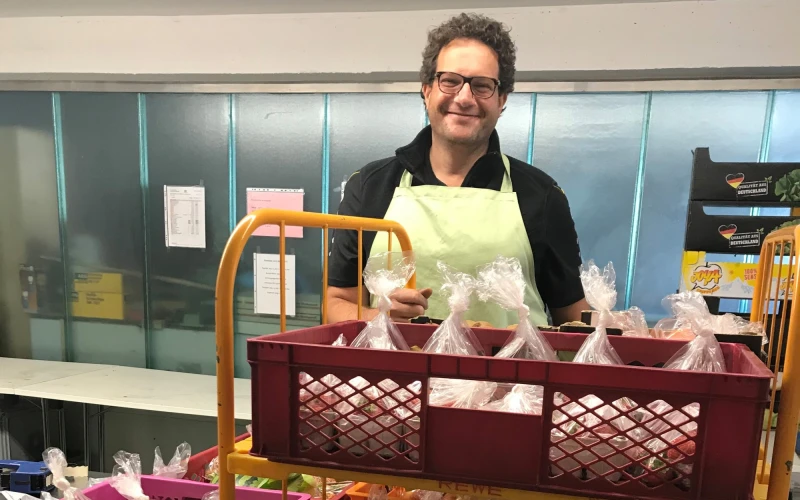 Ein Mann mit lockigen Haaren und Brille tr&auml;gt eine helle Sch&uuml;rze und steht hinter einem Wagen mit mehreren Kisten voller in Folie verpackter Lebensmittelportionen.