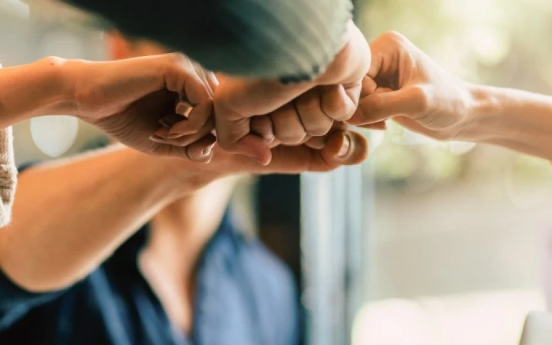 Mehrere H&auml;nde einer Gruppe von vier Menschen sto&szlig;en in einer Geste der Einheit und Zusammenarbeit mit den F&auml;usten zusammen, vor einem unscharfen Hintergrund in ged&auml;mpften Farbt&ouml;nen.