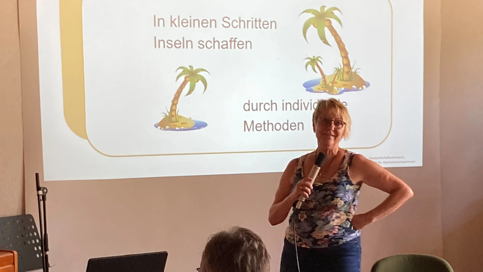 Eine Frau mit Mikrofon steht vor einer Pr&auml;sentationsfolie mit der Aufschrift "In kleinen Schritten Inseln schaffen", w&auml;hrend im Vordergrund der Kopf einer Person zu sehen ist.