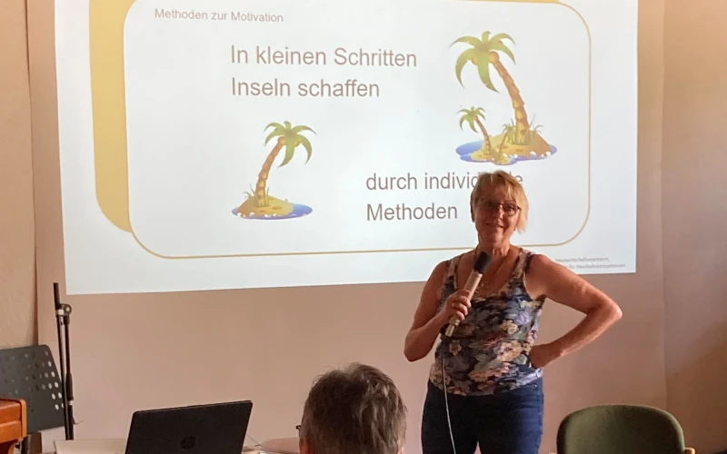 Eine Frau mit Mikrofon steht vor einer Pr&auml;sentationsfolie mit der Aufschrift "In kleinen Schritten Inseln schaffen", w&auml;hrend im Vordergrund der Kopf einer Person zu sehen ist.