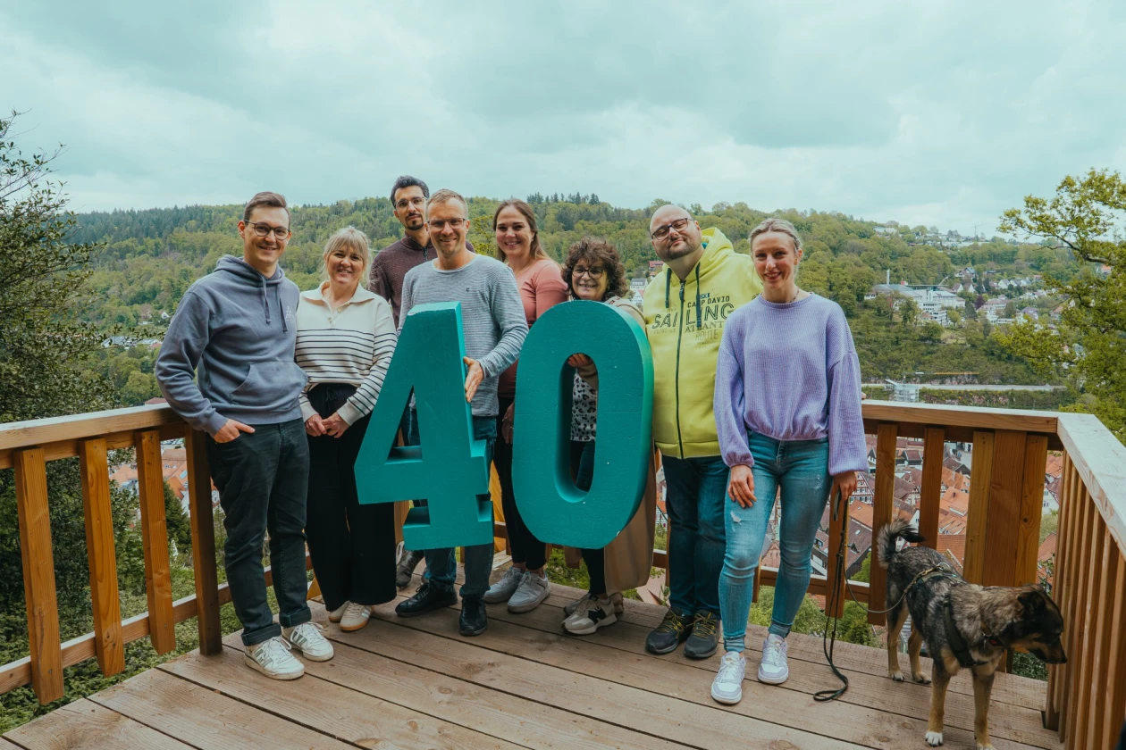 Einige Personen stehen auf einer Holzterrasse mit Aussicht auf eine gr&uuml;ne Landschaft und halten gro&szlig;e t&uuml;rkise Zahlen &bdquo;40" in die Kamera. Ein Hund steht rechts neben der Gruppe.