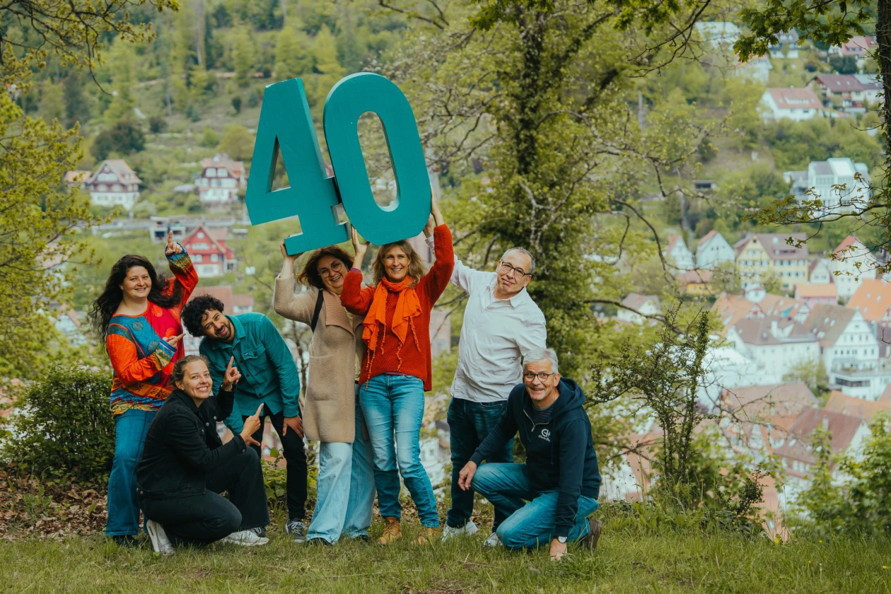 Eine Gruppe von Menschen h&auml;lt gemeinsam eine gro&szlig;e t&uuml;rkise Nummer 40 in die H&ouml;he und posiert auf einer Wiese mit B&auml;umen und einer Stadt im Hintergrund.