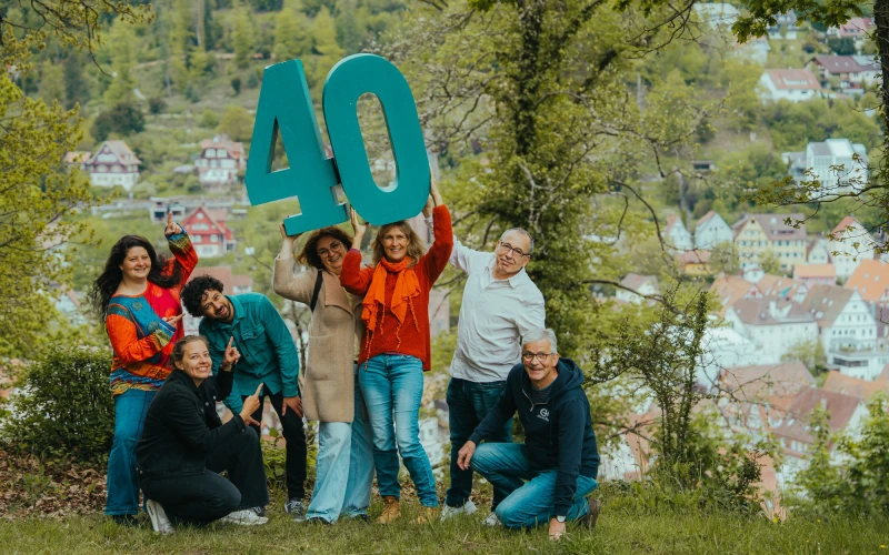 Eine Gruppe von Menschen h&auml;lt gemeinsam eine gro&szlig;e t&uuml;rkise Nummer 40 in die H&ouml;he und posiert auf einer Wiese mit B&auml;umen und einer Stadt im Hintergrund.