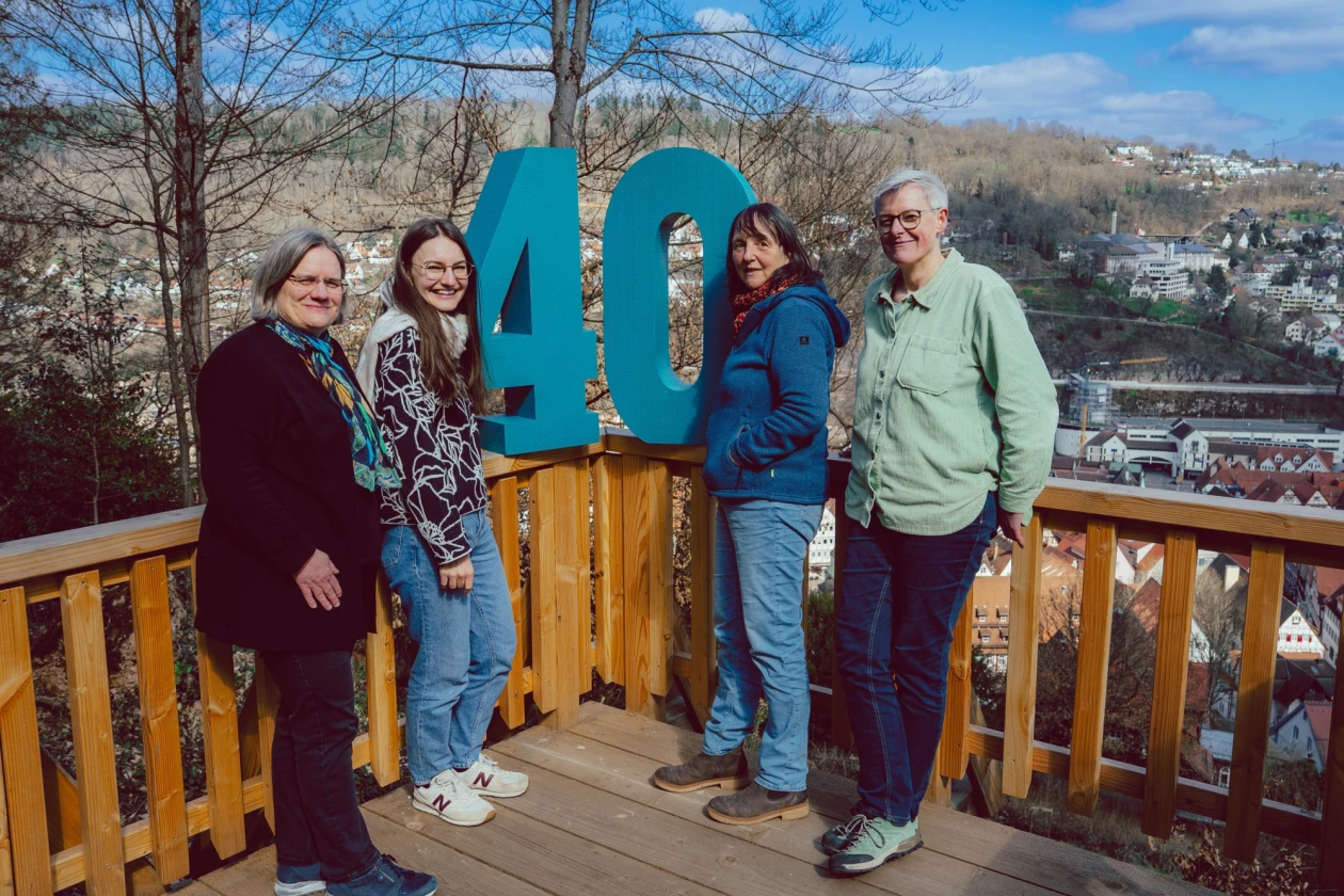 Vier Personen stehen auf einer Holzterrasse vor einer gro&szlig;en t&uuml;rkisen Nummer 40 und l&auml;cheln in die Kamera