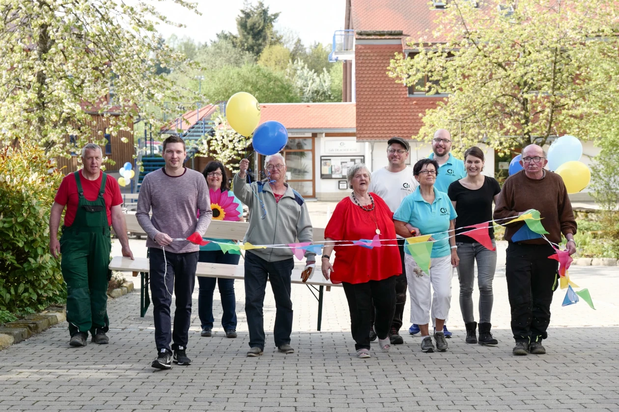 Gruppe von Menschen unterschiedlichen Alters und Geschlechts stehen mit bunten Luftballons und Wimpeln auf einem Pflasterweg. Im Hintergrund sind ein Geb&auml;ude mit blauer Treppe und B&auml;ume zu sehen.