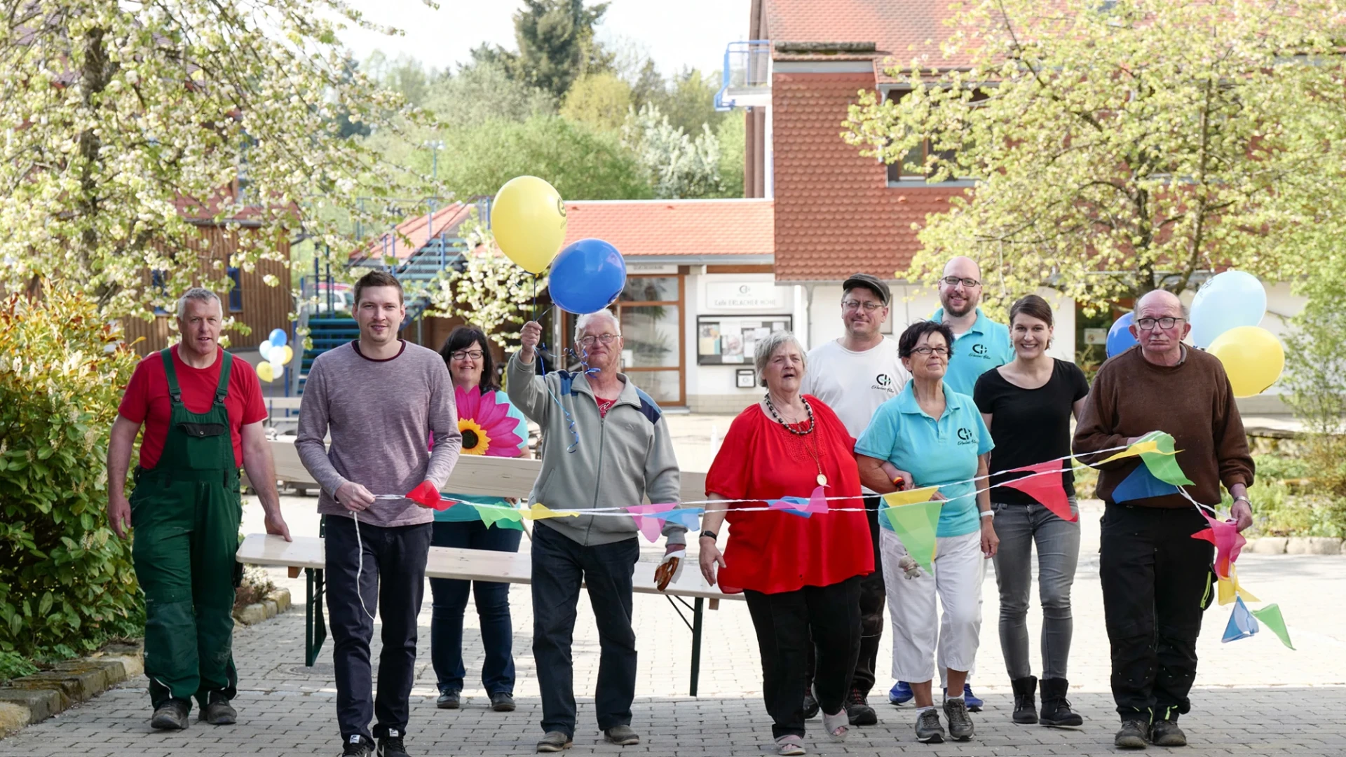 Gruppe von Menschen unterschiedlichen Alters und Geschlechts stehen mit bunten Luftballons und Wimpeln auf einem Pflasterweg. Im Hintergrund sind ein Geb&auml;ude mit blauer Treppe und B&auml;ume zu sehen.