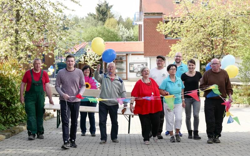 Gruppe von Menschen unterschiedlichen Alters und Geschlechts stehen mit bunten Luftballons und Wimpeln auf einem Pflasterweg. Im Hintergrund sind ein Geb&auml;ude mit blauer Treppe und B&auml;ume zu sehen.