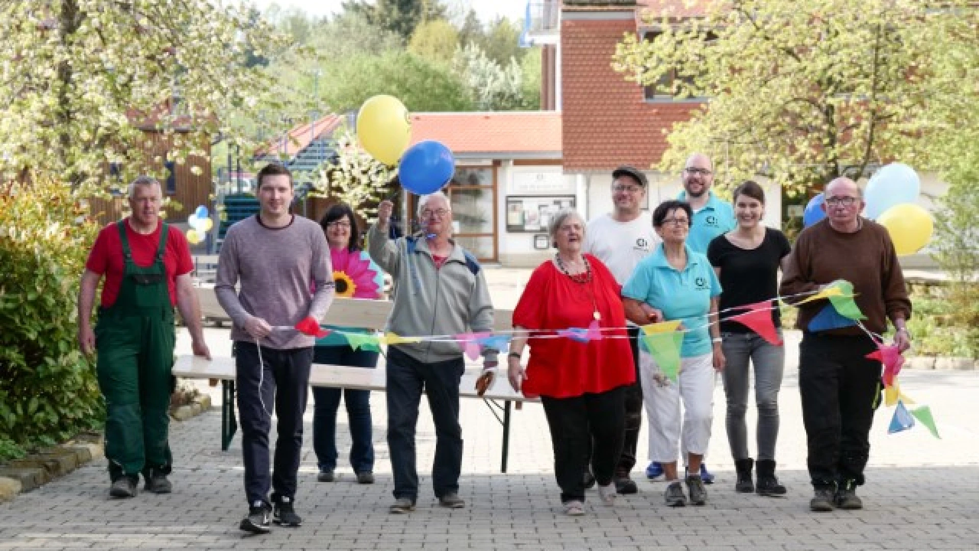 Gruppe von etwa zehn Menschen verschiedenen Alters beim Spaziergang auf einem Pflasterweg, einige tragen bunte Luftballons, sie scheinen gemeinsam an einer Veranstaltung oder Feier teilzunehmen.