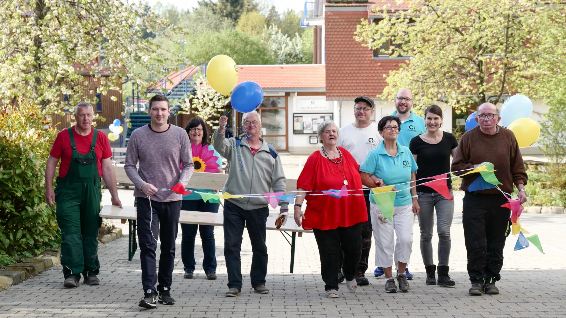 Gruppe von Menschen unterschiedlichen Alters und Geschlechts stehen mit bunten Luftballons und Wimpeln auf einem Pflasterweg. Im Hintergrund ist ein Geb&auml;ude mit blauer Treppe und B&auml;ume zu sehen.