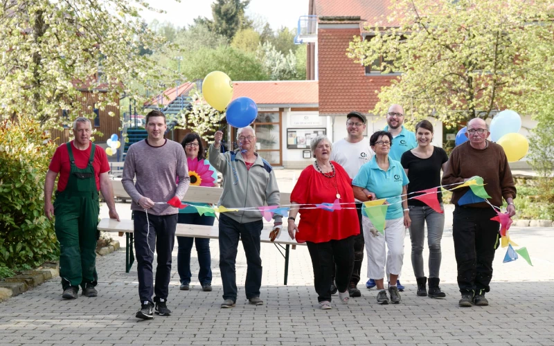 Gruppe von Menschen unterschiedlichen Alters und Geschlechts stehen mit bunten Luftballons und Wimpeln auf einem Pflasterweg. Im Hintergrund ist ein Geb&auml;ude mit blauer Treppe und B&auml;ume zu sehen.