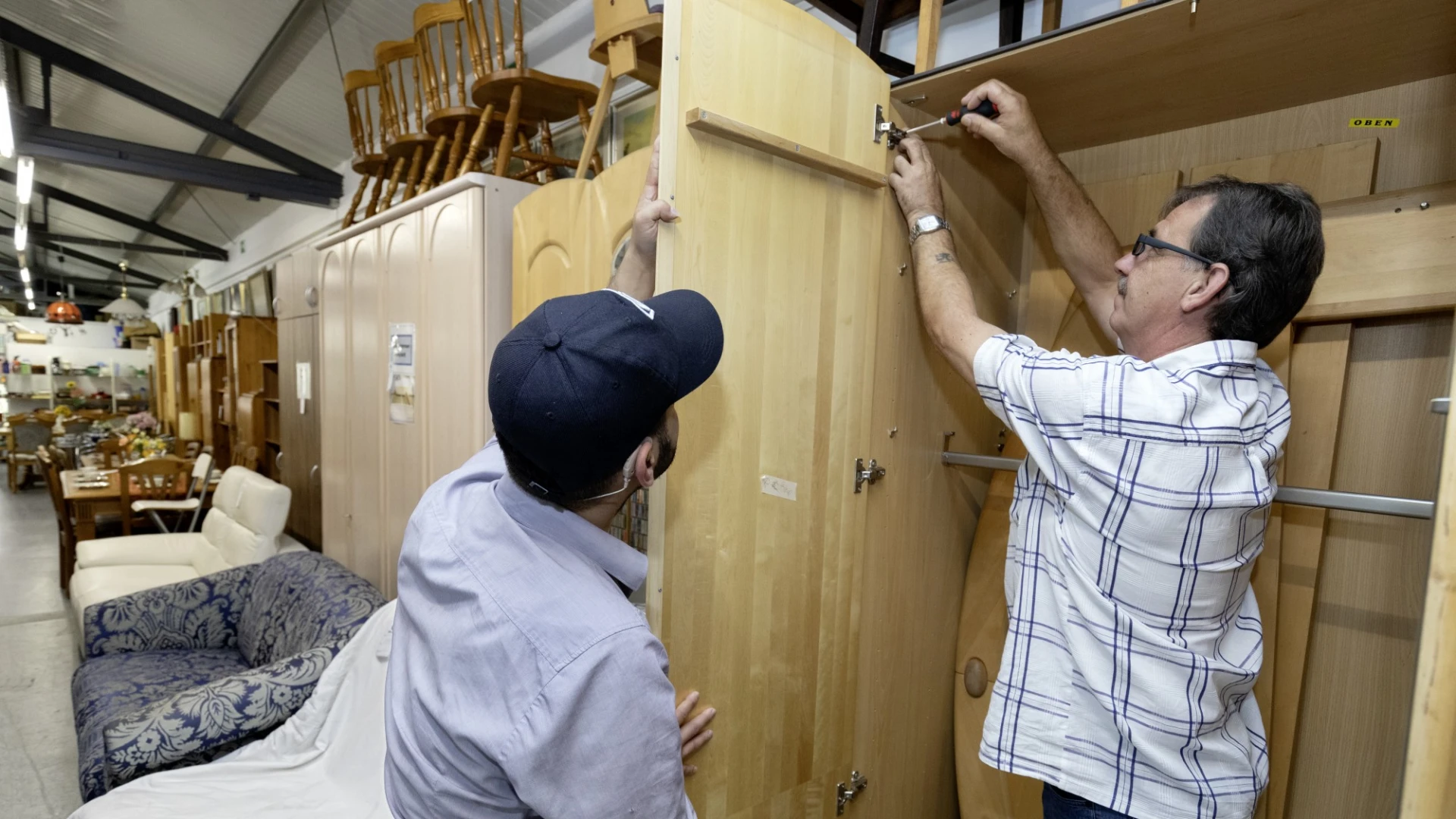 Ein Mann mit einer Schildm&uuml;tze h&auml;lt eine Schrankt&uuml;r, w&auml;hrend ein anderer Mann diese mit einem Schraubendreher am Schrank befestigt. Im Hintergrund sind weitere Schr&auml;nke und M&ouml;bel zu sehen.