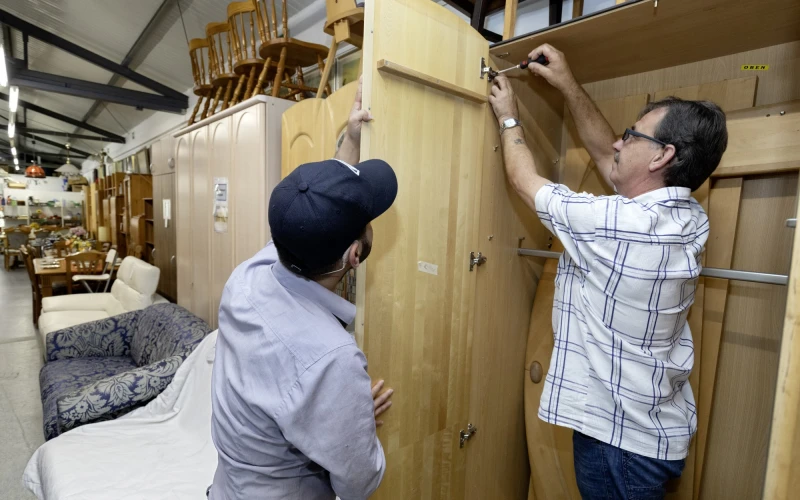 Ein Mann mit einer Schildm&uuml;tze h&auml;lt eine Schrankt&uuml;r, w&auml;hrend ein anderer Mann diese mit einem Schraubendreher am Schrank befestigt. Im Hintergrund sind weitere Schr&auml;nke und M&ouml;bel zu sehen.