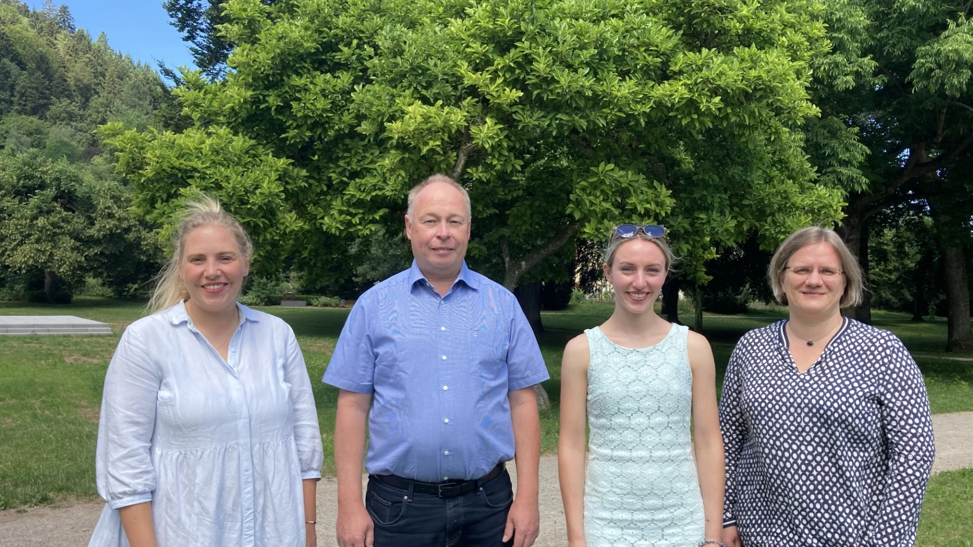 Drei j&uuml;ngere Frauen und ein Mann mittleren Alters stehen auf einer Wiese vor einem Hintergrund aus B&auml;umen. Alle l&auml;cheln freundlich in die Kamera.