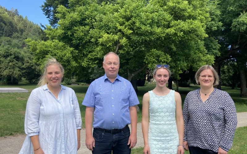 Drei j&uuml;ngere Frauen und ein Mann mittleren Alters stehen auf einer Wiese vor einem Hintergrund aus B&auml;umen. Alle l&auml;cheln freundlich in die Kamera.
