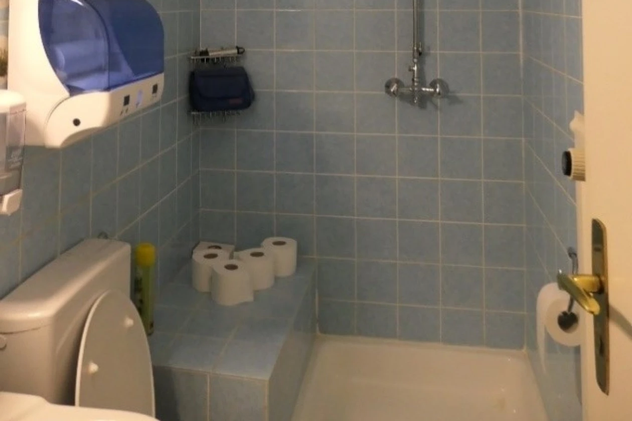 Ein Badezimmer mit blauen Wandfliesen ist zu sehen. Es gibt eine Dusche mit Vorhangstange, eine Toilette und mehrere Toilettenpapierrollen.