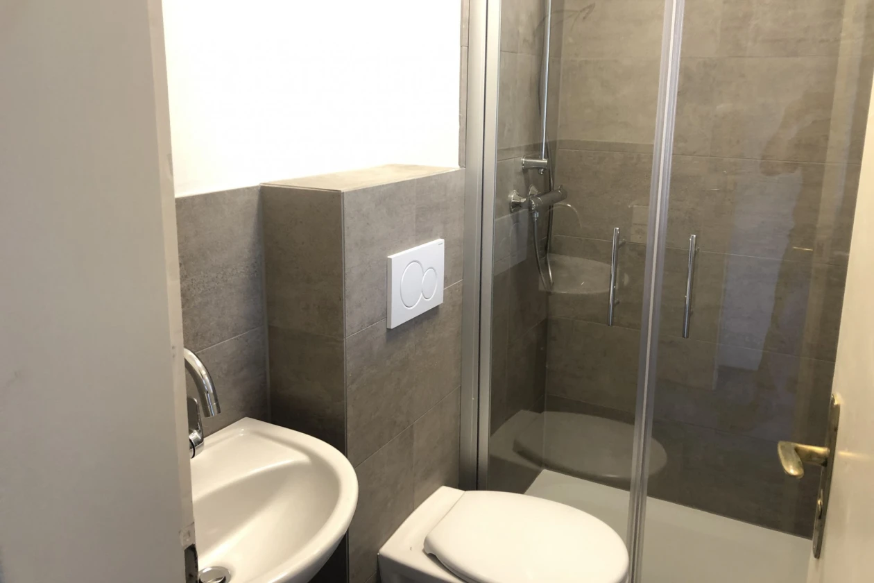 Ein Bad mit Waschbecken, Wandtoilette und einer Dusche mit Glast&uuml;r ist zu sehen.