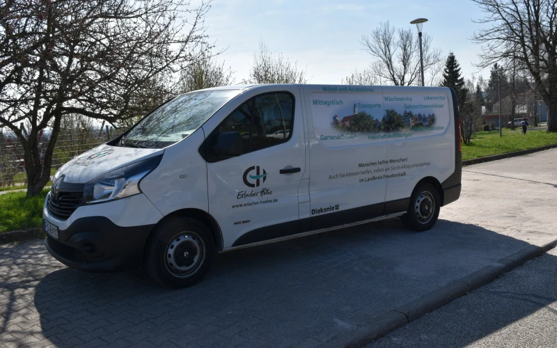 Ein wei&szlig;er Lieferwagen mit dem Logo der ErlacherH&ouml;he" steht auf einem Parkplatz neben kahlen B&auml;umen und gr&uuml;nen Wiesen.