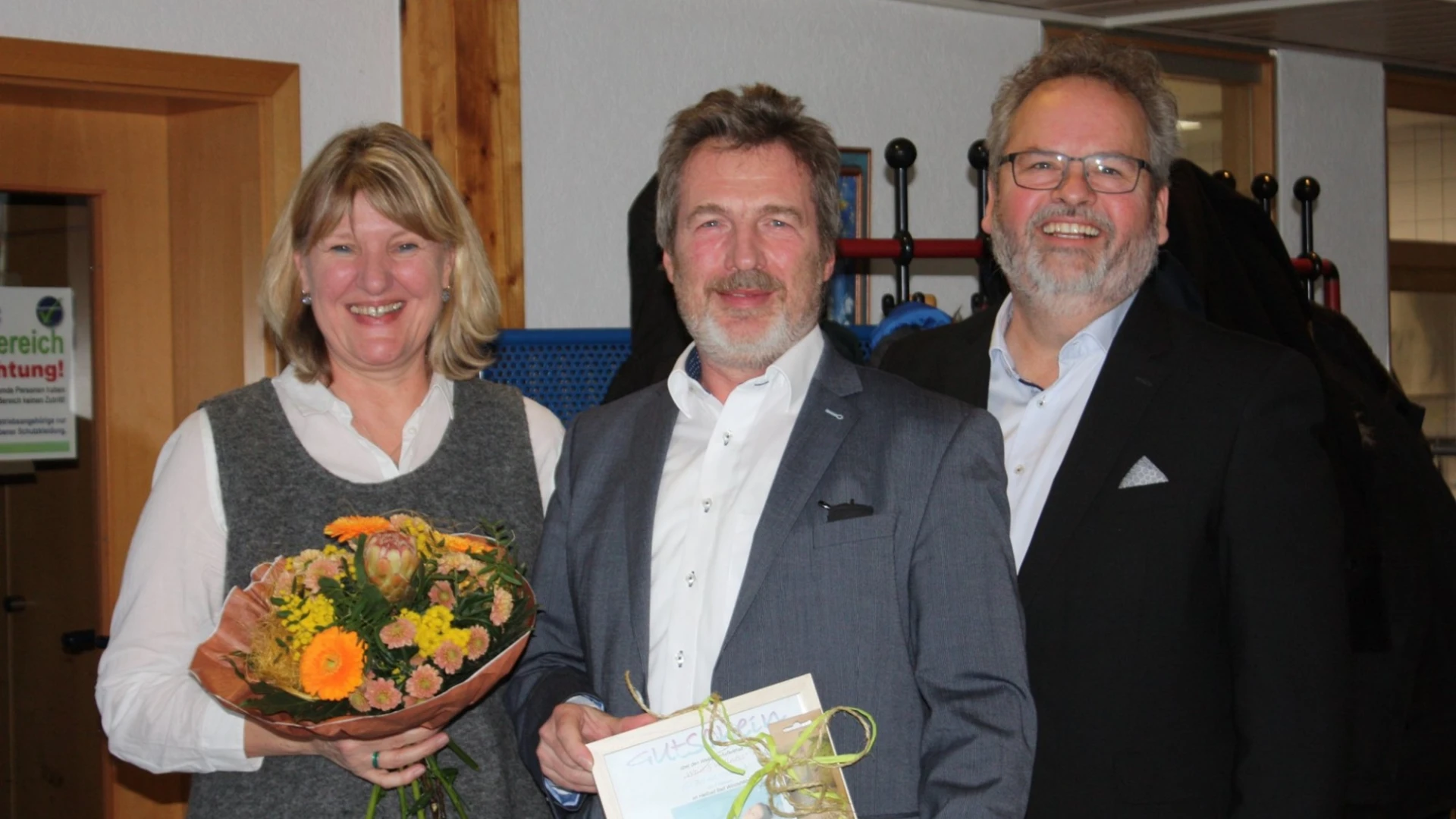 Drei Personen stehen nebeneinander und lachen in die Kamera. Die Frau links h&auml;lt einen Blumenstrau&szlig; in der Hand. Daneben stehen zwei M&auml;nner im Anzug. Der Mann in der Mitte h&auml;lt einen Geschenk mit einer Schleife in der Hand. Im Hintergrund ist ein T&uuml;rrahmen, eine Wand und ein Garderobenst&auml;nder zu sehen.