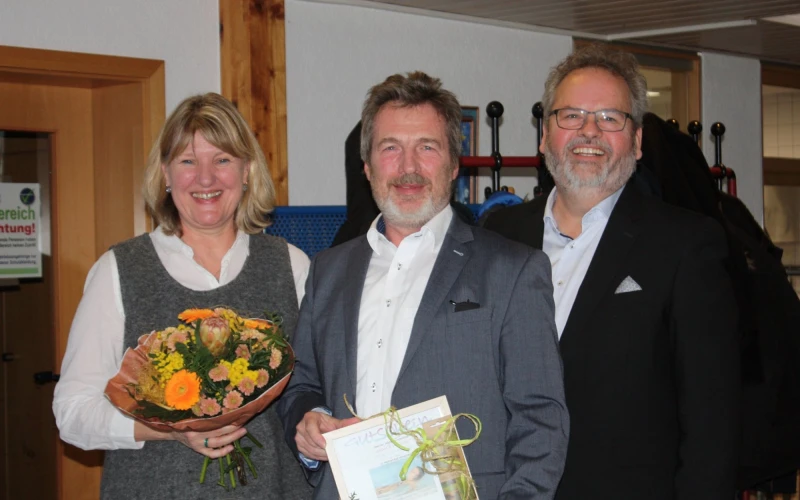 Drei Personen stehen nebeneinander und lachen in die Kamera. Die Frau links h&auml;lt einen Blumenstrau&szlig; in der Hand. Daneben stehen zwei M&auml;nner im Anzug. Der Mann in der Mitte h&auml;lt einen Geschenk mit einer Schleife in der Hand. Im Hintergrund ist ein T&uuml;rrahmen, eine Wand und ein Garderobenst&auml;nder zu sehen.