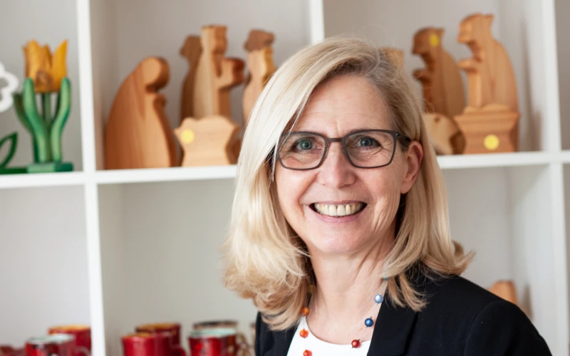 L&auml;chelnde Frau mit schulterlangem blondem Haar und Brille steht vor einem Regal mit dekorativen Holzfiguren. Sie tr&auml;gt eine bunte Perlenhalskette.